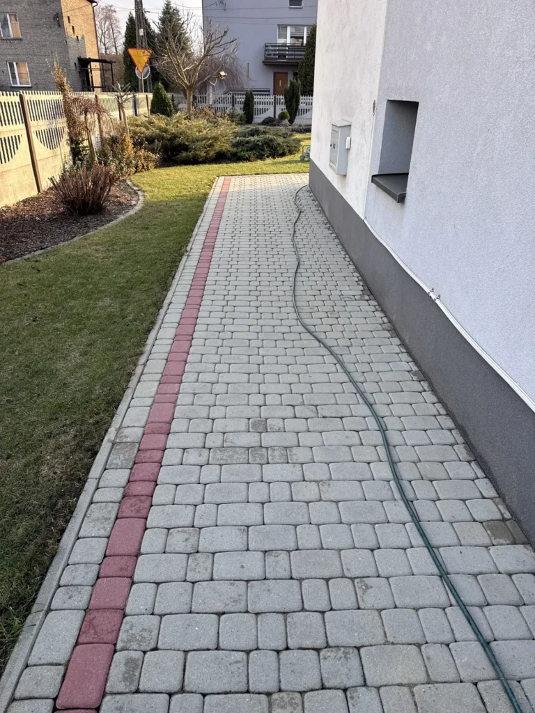 Przygotowanie frontu robót do gruntownego zmywania kostki brukowej prowadzącej wzdłuż elewacji domu mieszkalnego.