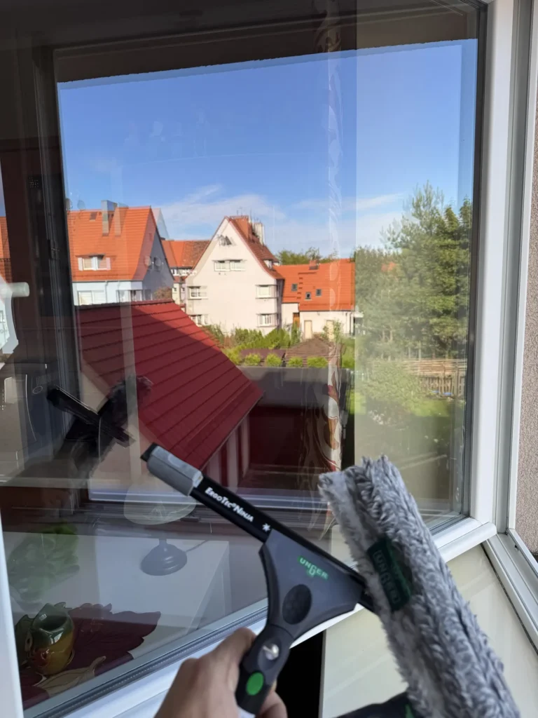 Szyba okienna myta od zewnątrz profesjonalną ściągaczką do szyb z widokiem na duże drzewa iglaste.